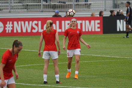 jogadora de futebol com camisa vermelha, calões e meias brancas trocando passes de cabeça com companheira de time em treino